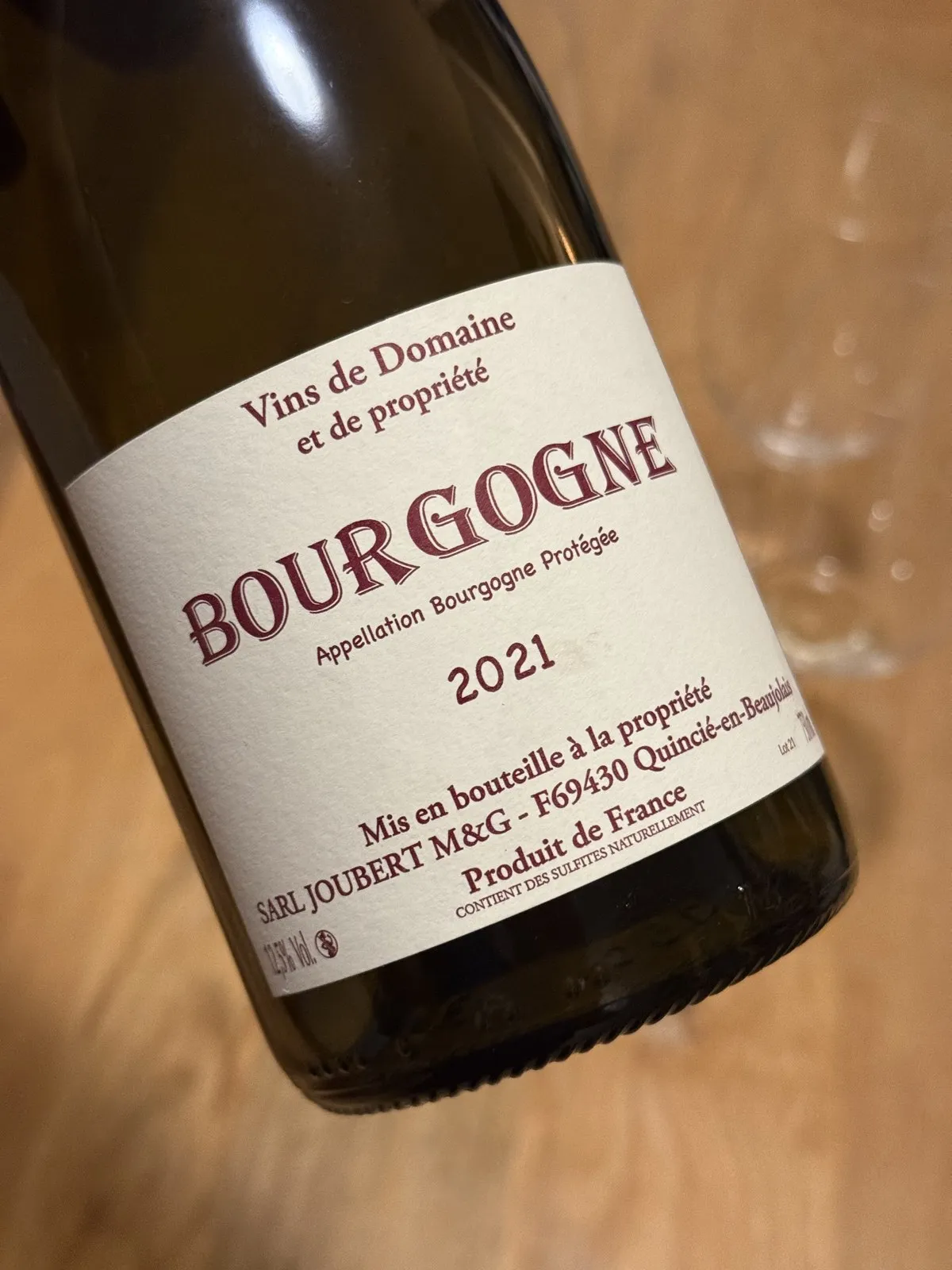 Primer plano de la etiqueta del Bourgogne Blanc 2021 de Domaine Joubert, con letras granate sobre papel crema, la botella inclinada sobre una mesa de madera y una copa vacía desenfocada al fondo.