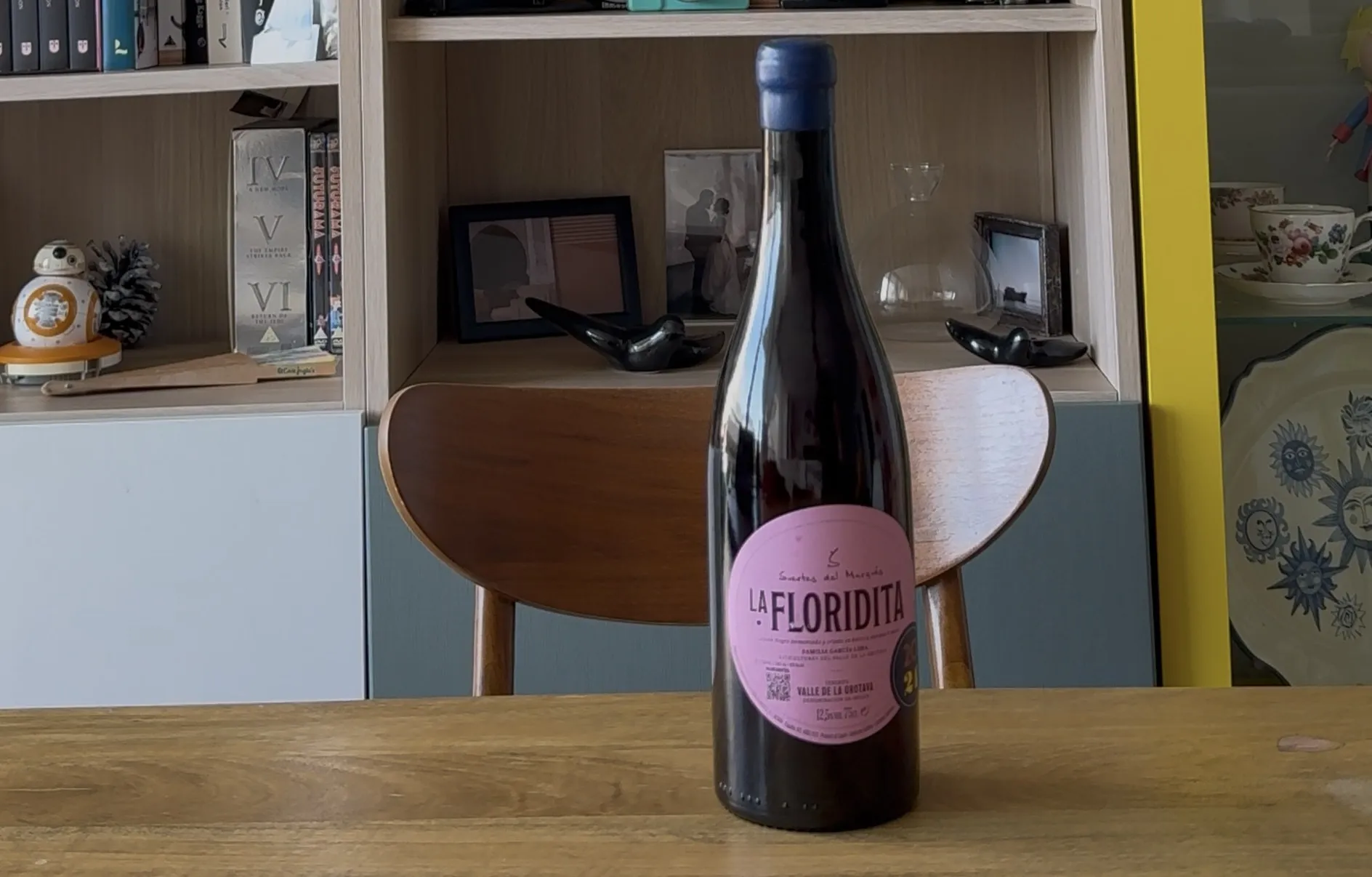 Botella de La Floridita 2024 de Suertes del Marqués, Listán Negro de la DO Valle de La Orotava, con su etiqueta rosa y cápsula de lacre azul sobre una mesa de madera, delante de una silla y una estantería.