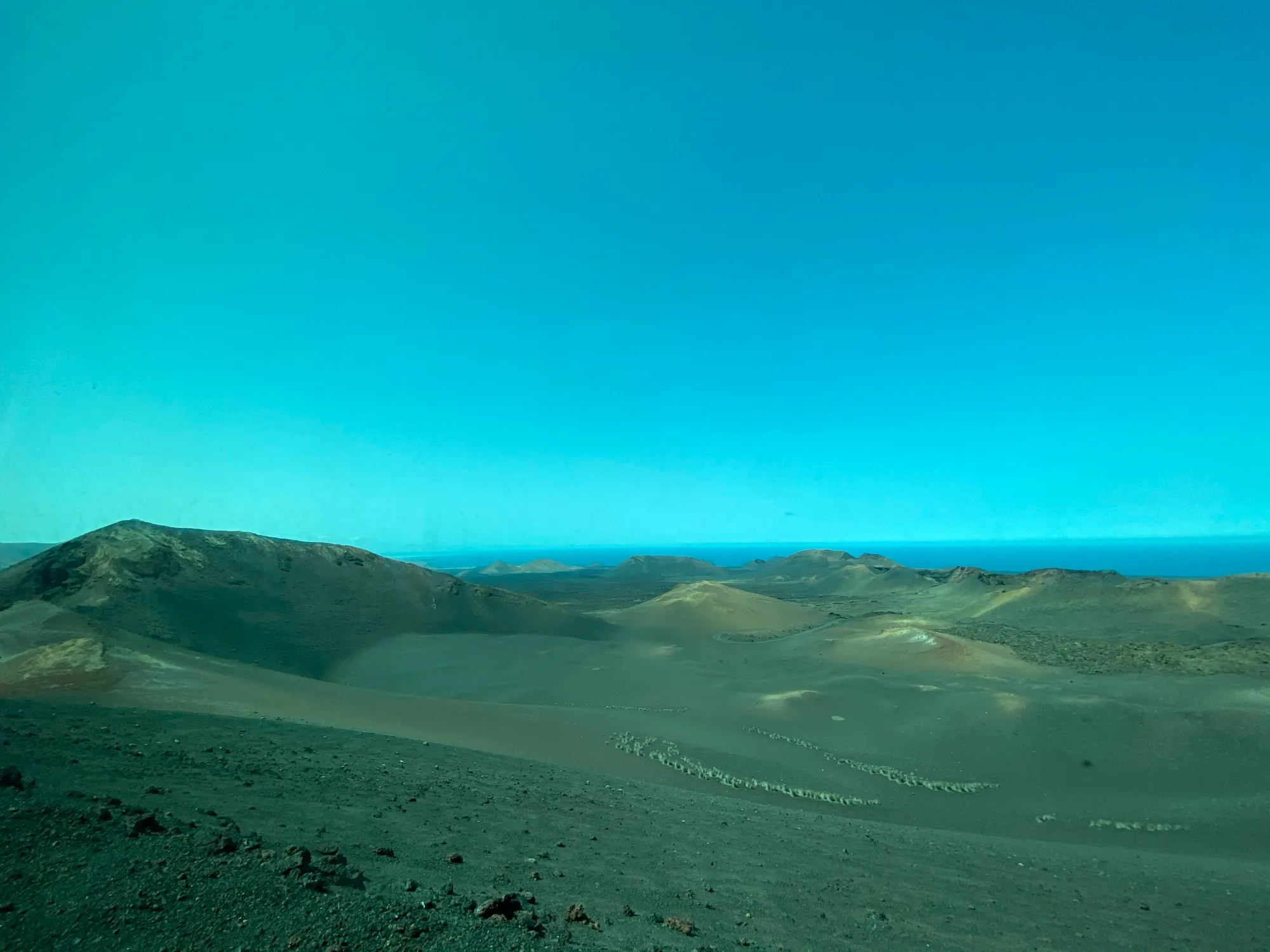 Paisaje volcánico del Parque Nacional de Timanfaya, en Lanzarote: conos y cráteres de ceniza y lapilli sobre un desierto negro y rojizo, con el Atlántico al fondo bajo un cielo despejado.