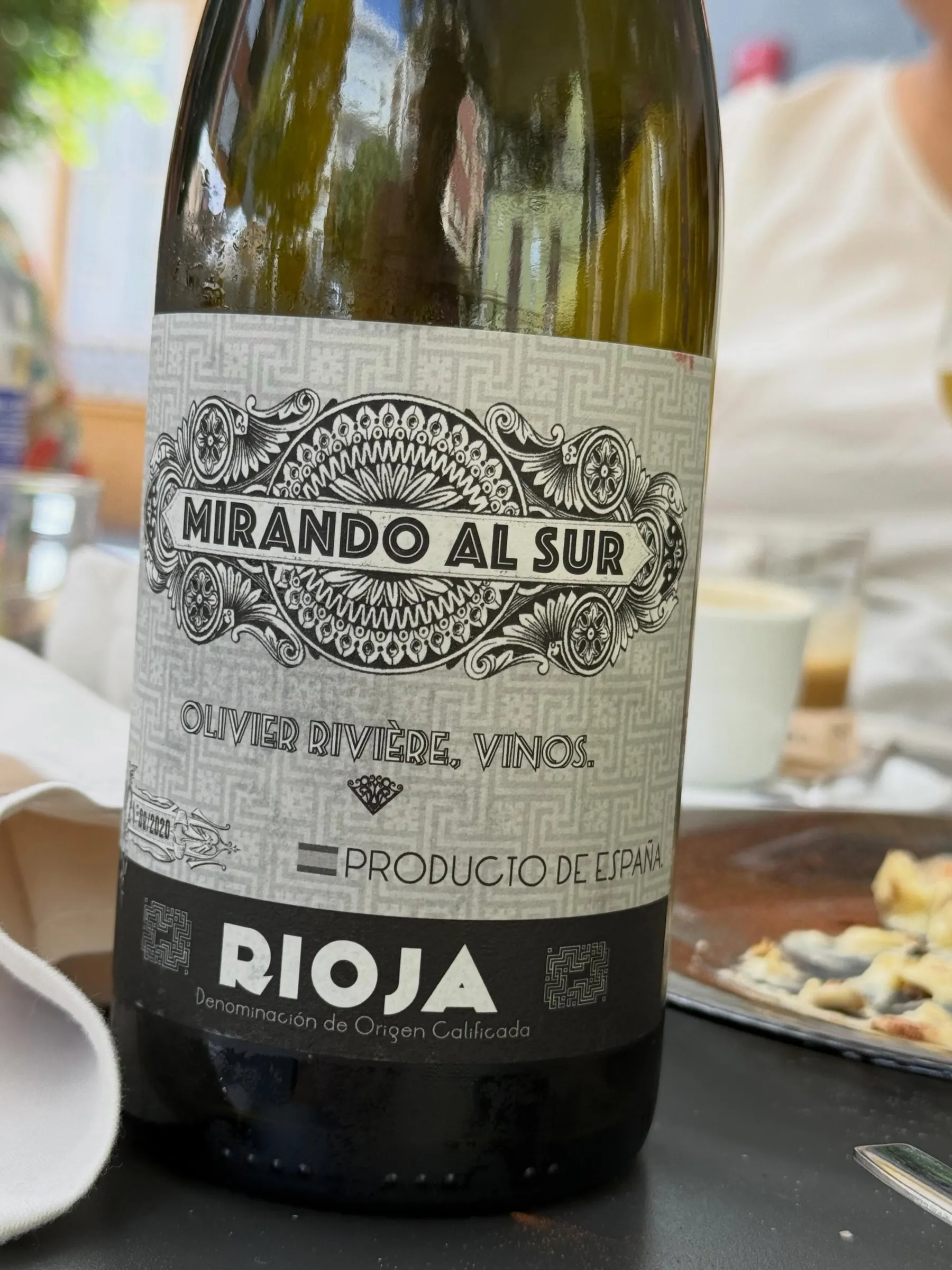 Botella de Mirando al Sur 2020, blanco de Viura de Olivier Rivière con DOCa Rioja, con su etiqueta gris de grecas y letras negras, en una mesa de terraza al sol junto a un plato y una taza de café.