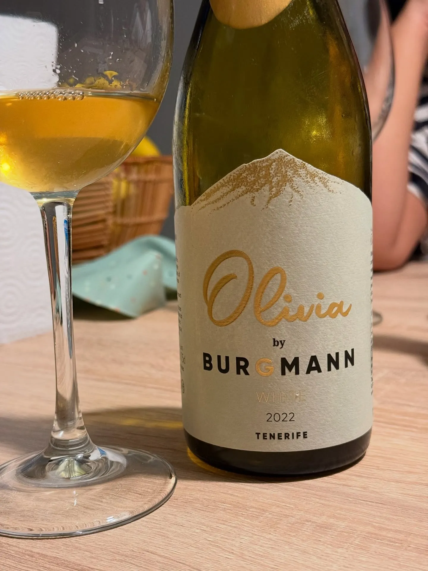 Botella del Olivia 2022 de Winery Burgmann, listán blanco de Tacoronte en Tenerife, con su etiqueta crema y el nombre en letra dorada, junto a una copa de vino blanco de color dorado intenso sobre una mesa de madera.